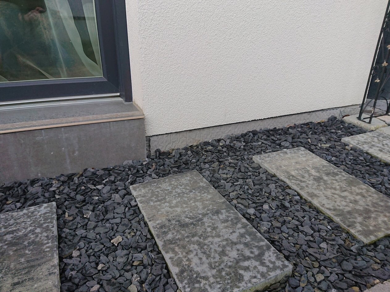 Außenansicht: Glas-Tür links, beige Wand, schwarzer Kiesweg mit rechteckigen Steinplatten.