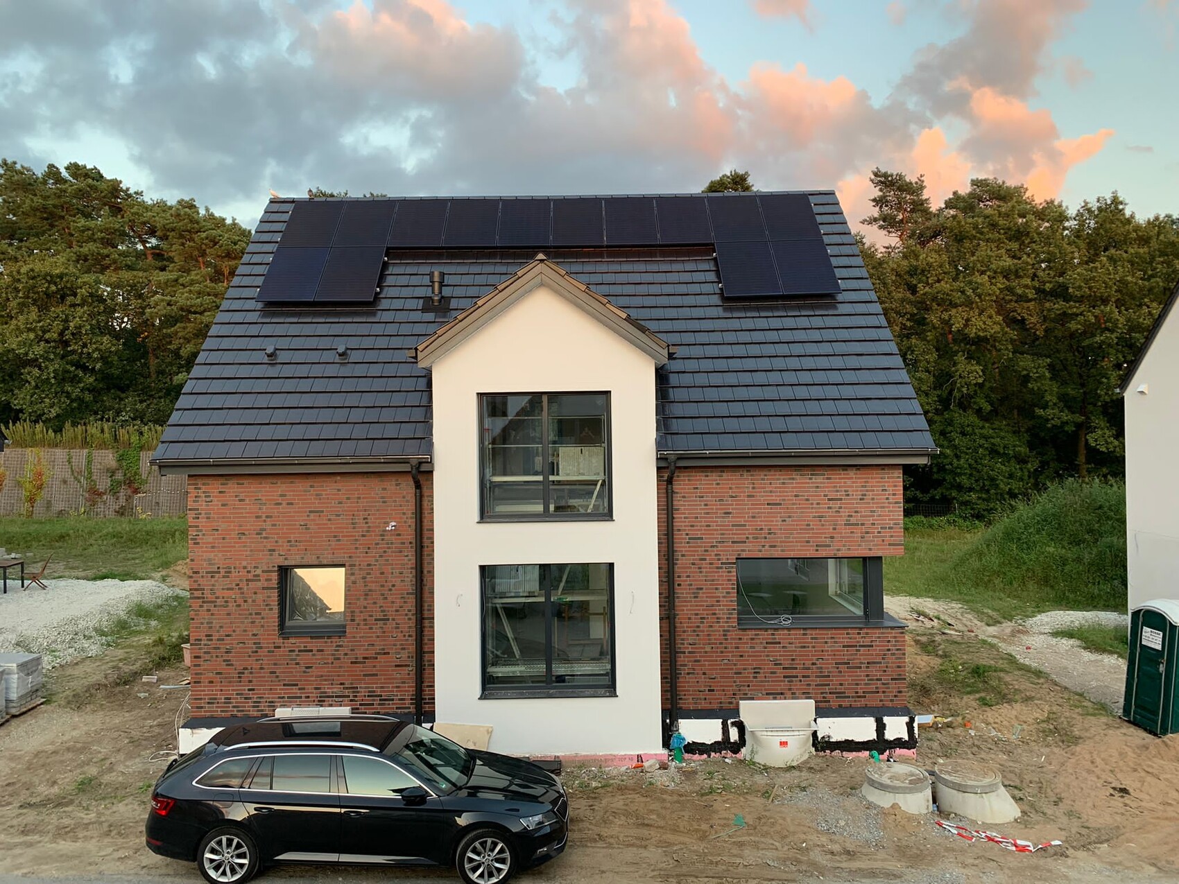 Zweistöckiges Haus mit dunklem Ziegeldach, Solarzellen, rotem Backstein und schwarzem Auto davor.
