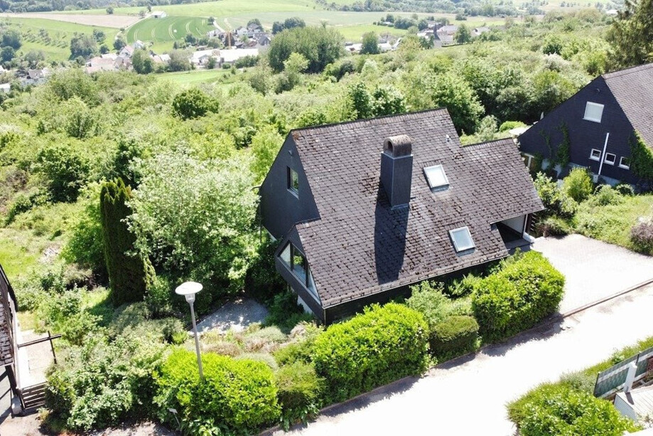 Luftaufnahme: Dunkles Einfamilienhaus mit Kamin, Dachfenstern, umgeben von grünen Hecken und Garten.