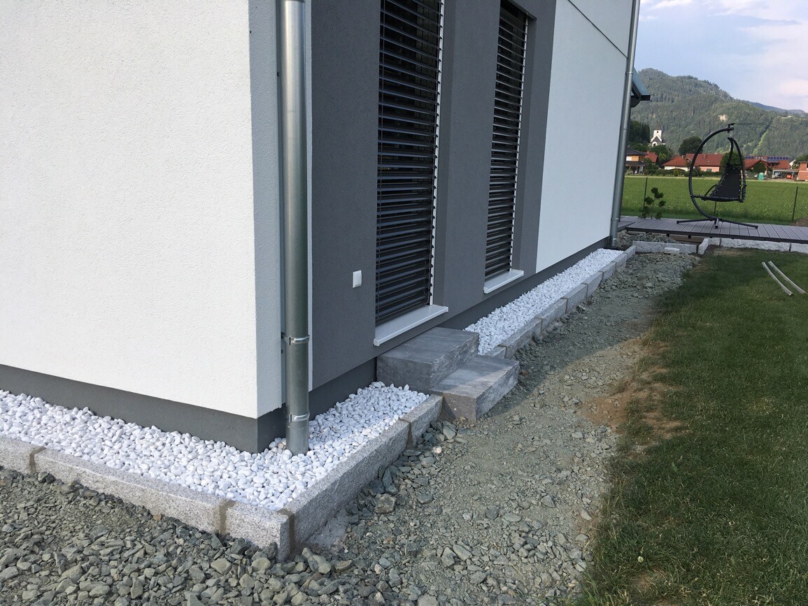 Aussenansicht eines modernen Hauses mit schwarzen Rolllaeden, Kiesbelag und Garten