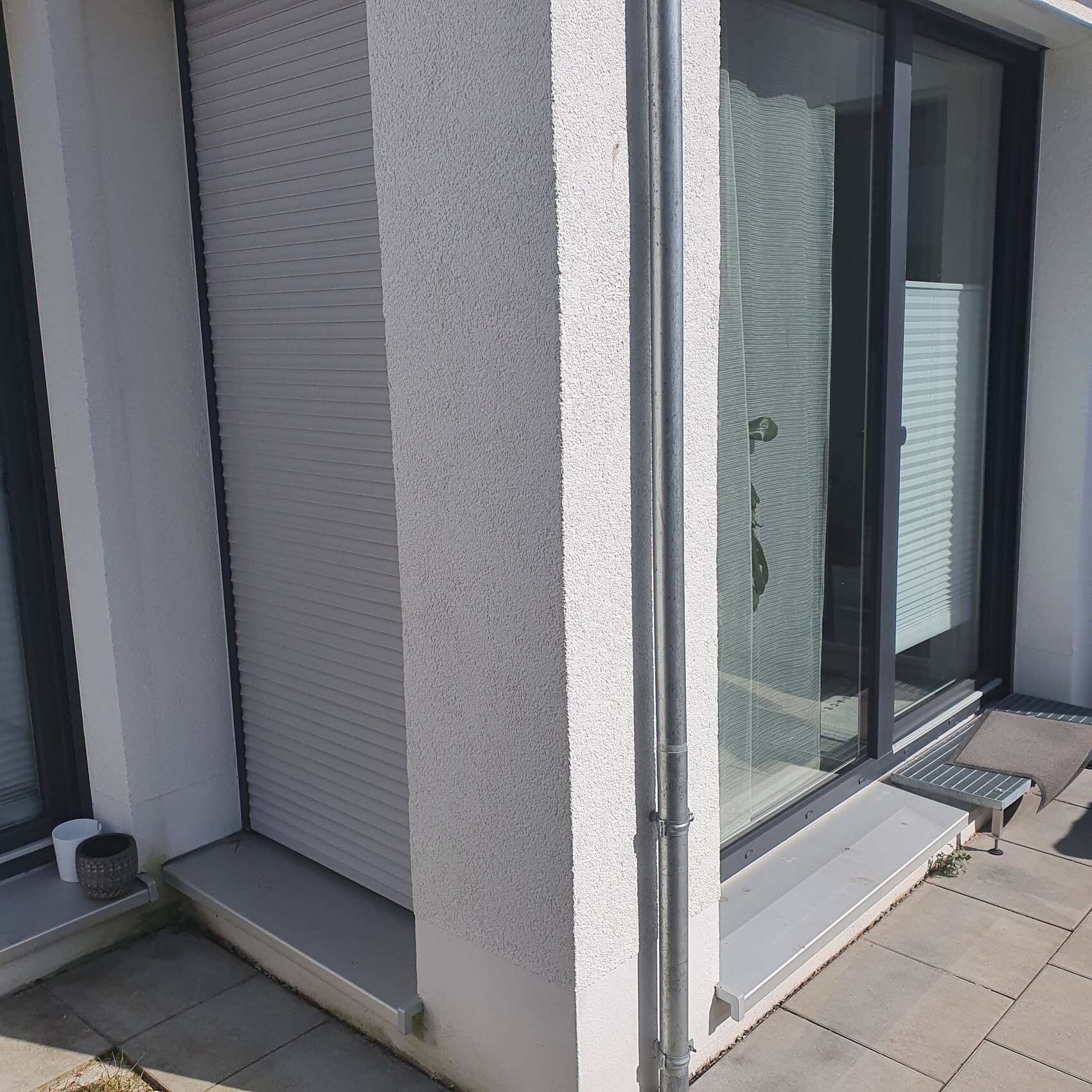 Weiße Fassade mit Fallrohr, große Glasfront mit Rollläden und Terrasse.