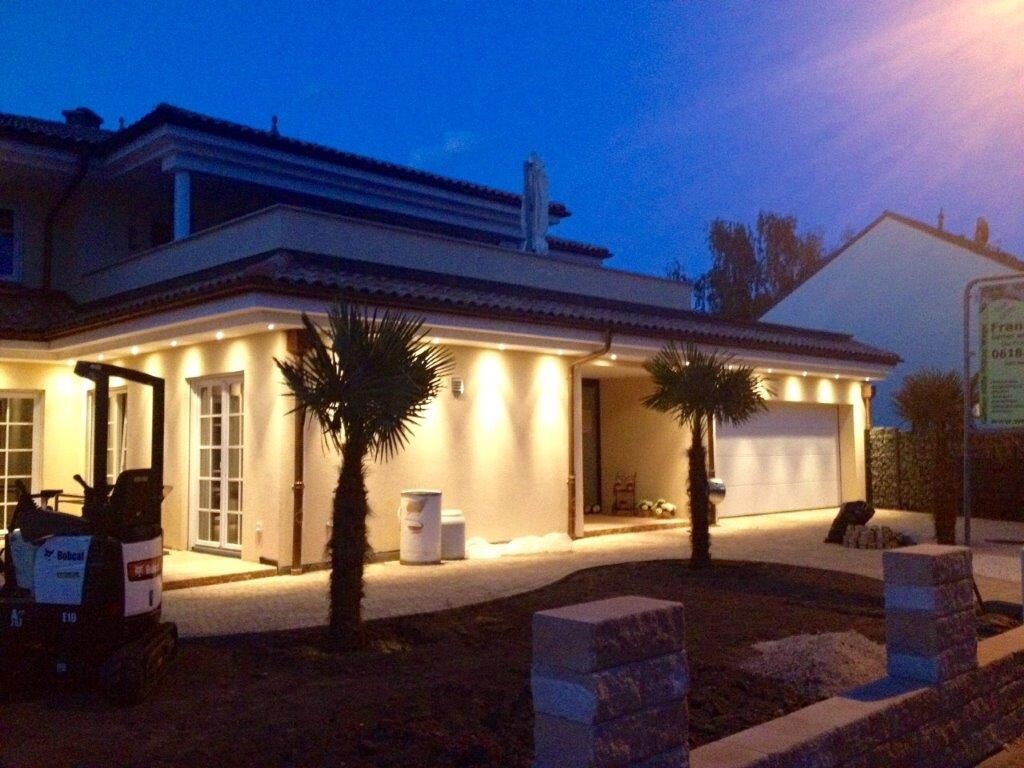 Moderne Einfamilienhausfront mit beleuchteter Fassade, Palmen, Garage und Zufahrt bei Abenddämmerung.