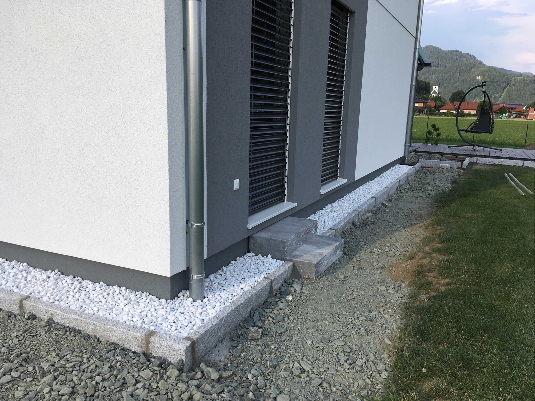 Moderne Hausfassade mit grauen Rollläden, Kiesstreifen und Steinstufen am Rand.