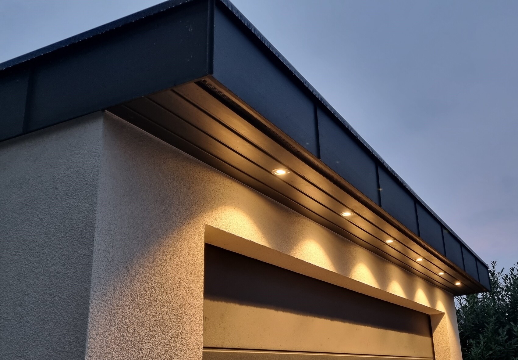 Moderne Garage mit dunklem Vordach und eingebauten Lampen; warme Lichtkegel an der Wand.