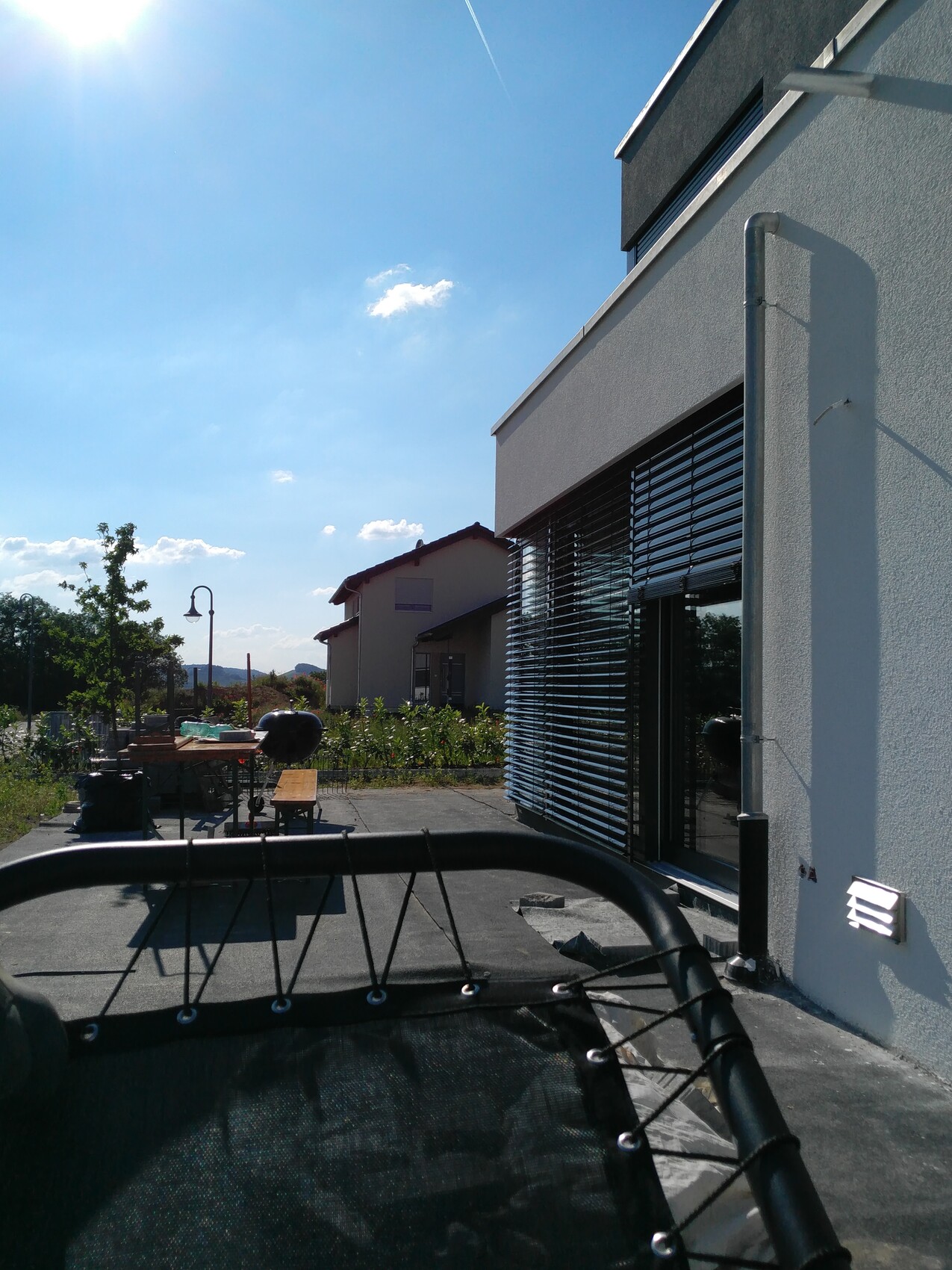 Moderne Hausfront mit großen Fenstern und Rollläden, Grill und Gartenmöbel im sonnigen Hof.