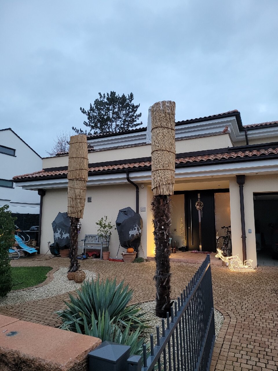 Vorderseite eines modernen Hauses mit zwei Palmenstämme in Strohdekoration, beleuchteter Eingang.
