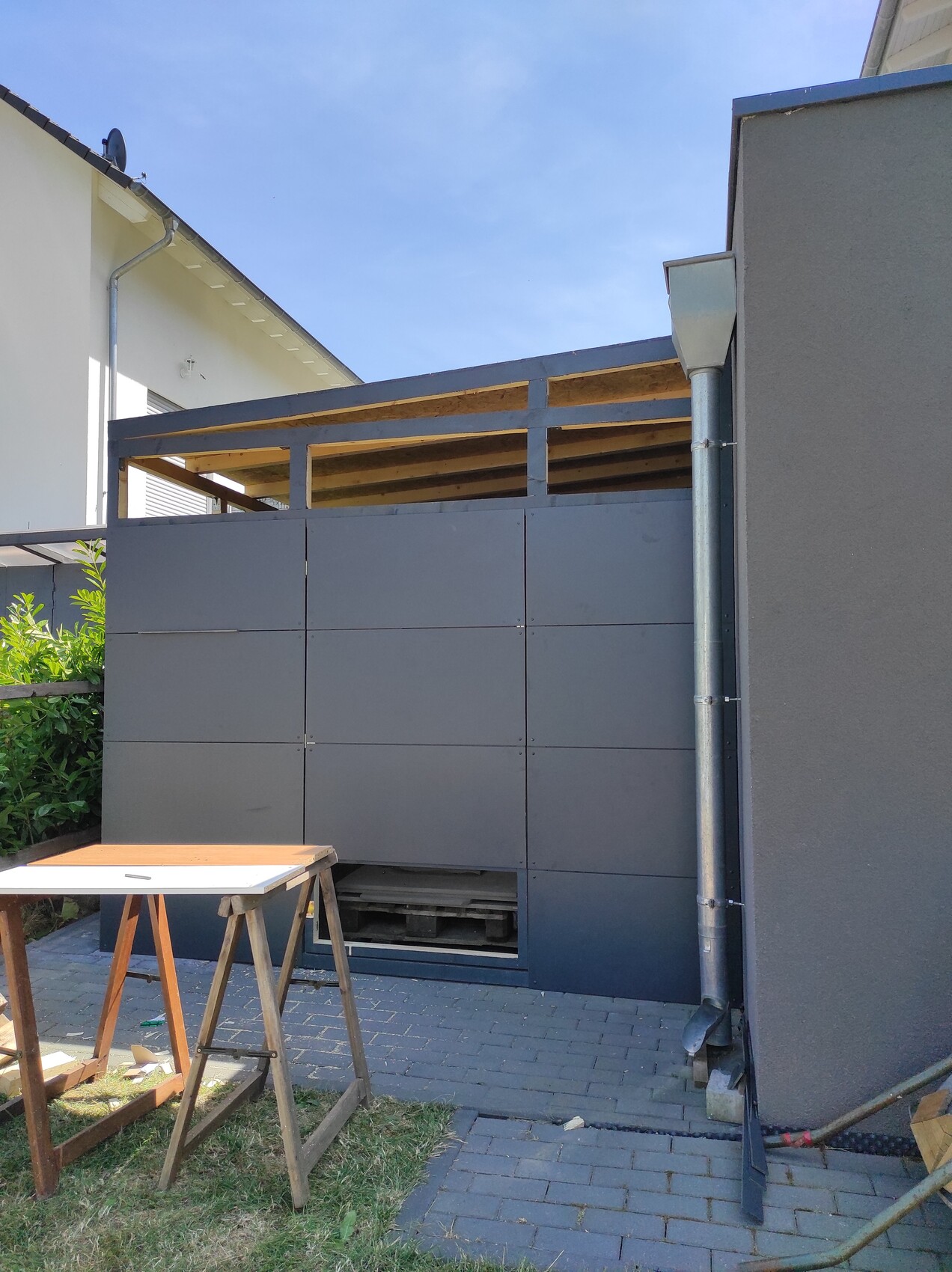 Außenansicht: graue, quadratische Metallwand mit offener Oberkante; Bau-Tisch und Holzlatten davor.