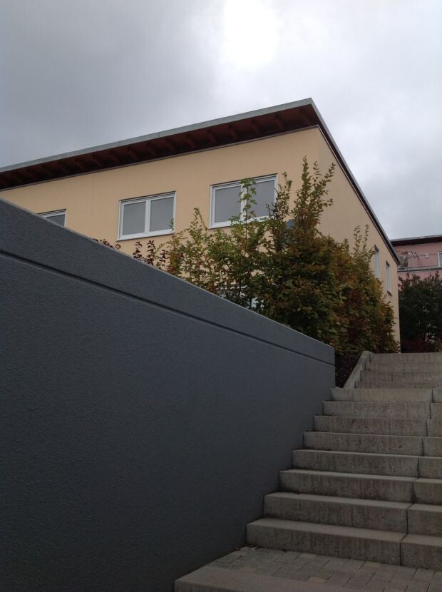Außenansicht eines beige gestrichenen Hauses mit Treppenaufgang und grauer Mauer.