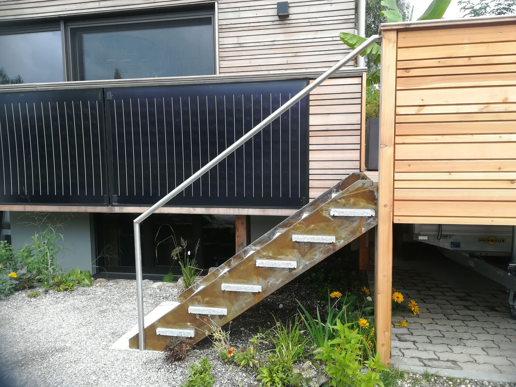 Außenansicht eines Hauses: Metalltreppe führt zur Holzterrasse, davor Pflanzen.