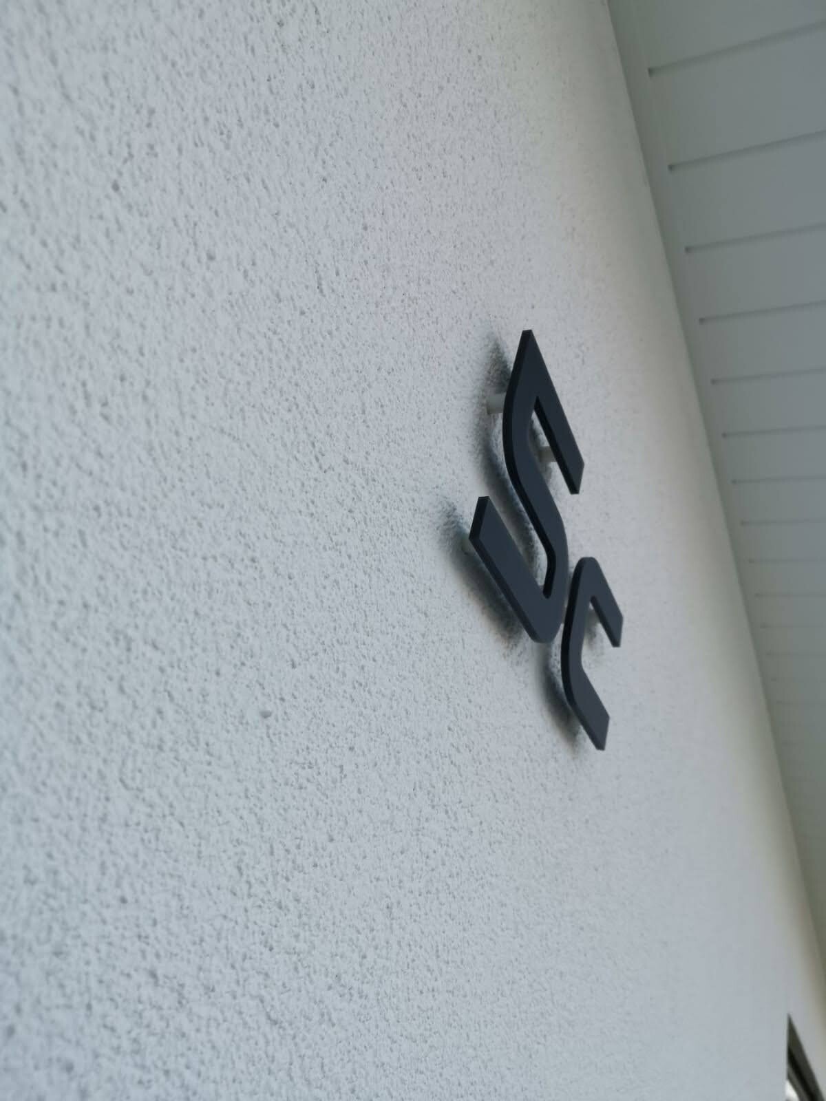 Dreidimensionales schwarzes Logo aus zwei stilisierten Buchstaben S und G an rauer weißer Wand.