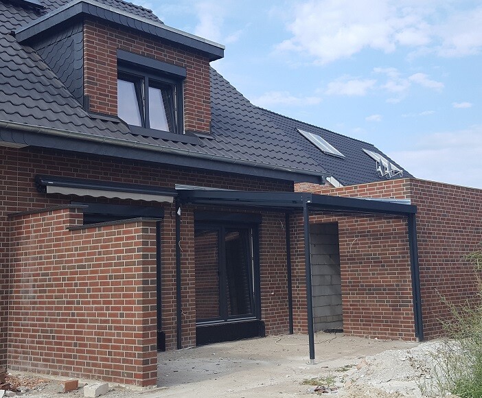 Zweistöckiges rotes Backsteinhaus mit dunklem Satteldach und offener Metallpergola im Bau.