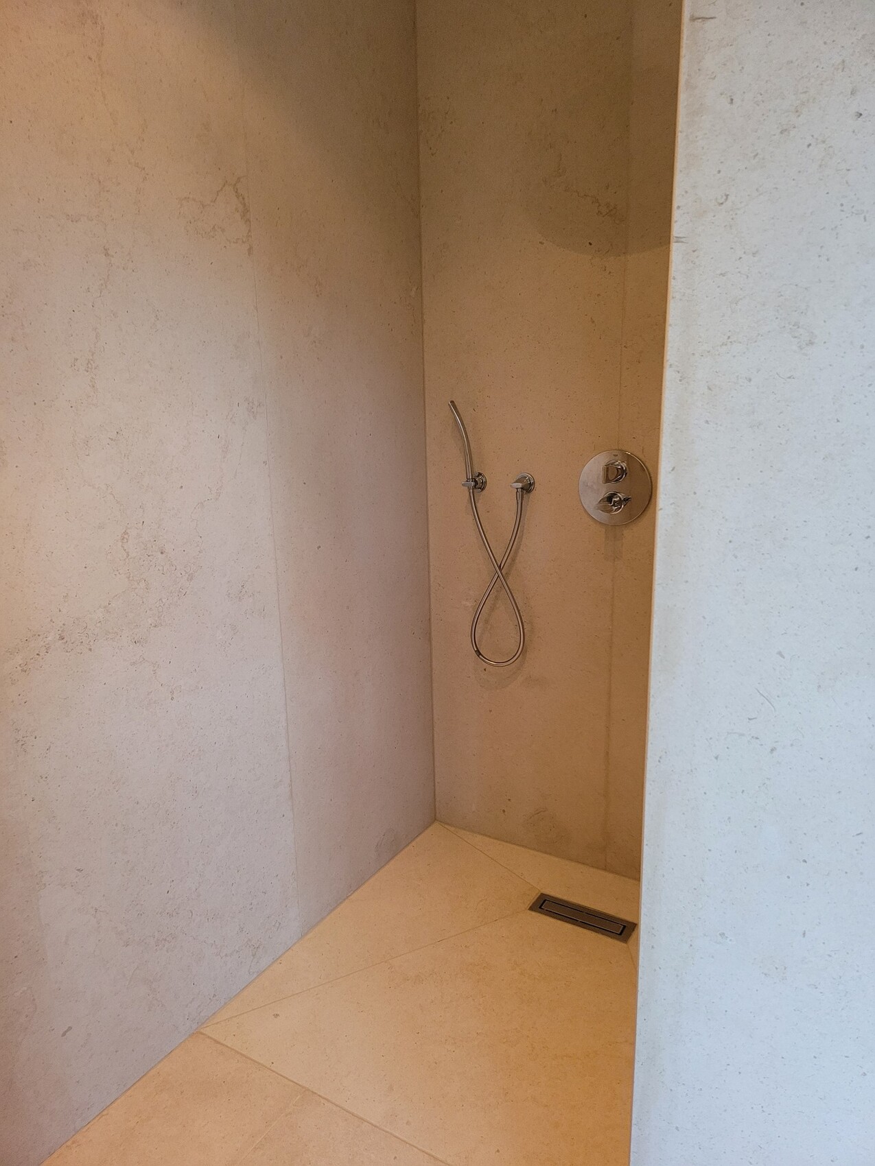 Dusche aus beigen Steinplatten mit einem flexiblen, schwenkbaren Brauseschlauch und Bodenablauf.