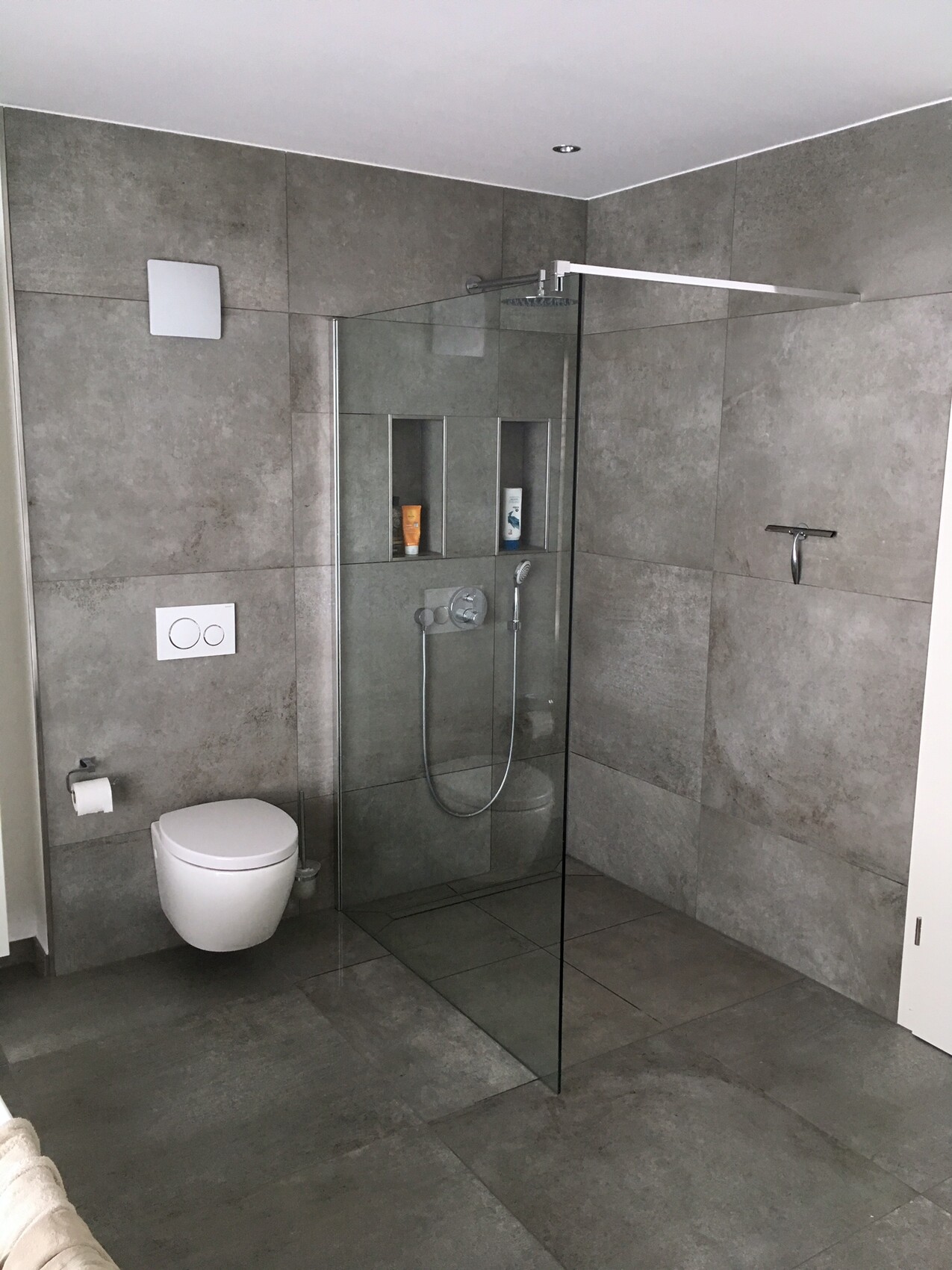 Modernes Badezimmer mit grauen Fliesen, Glasdusche, Wand-WC und Nischen mit Duschprodukten.