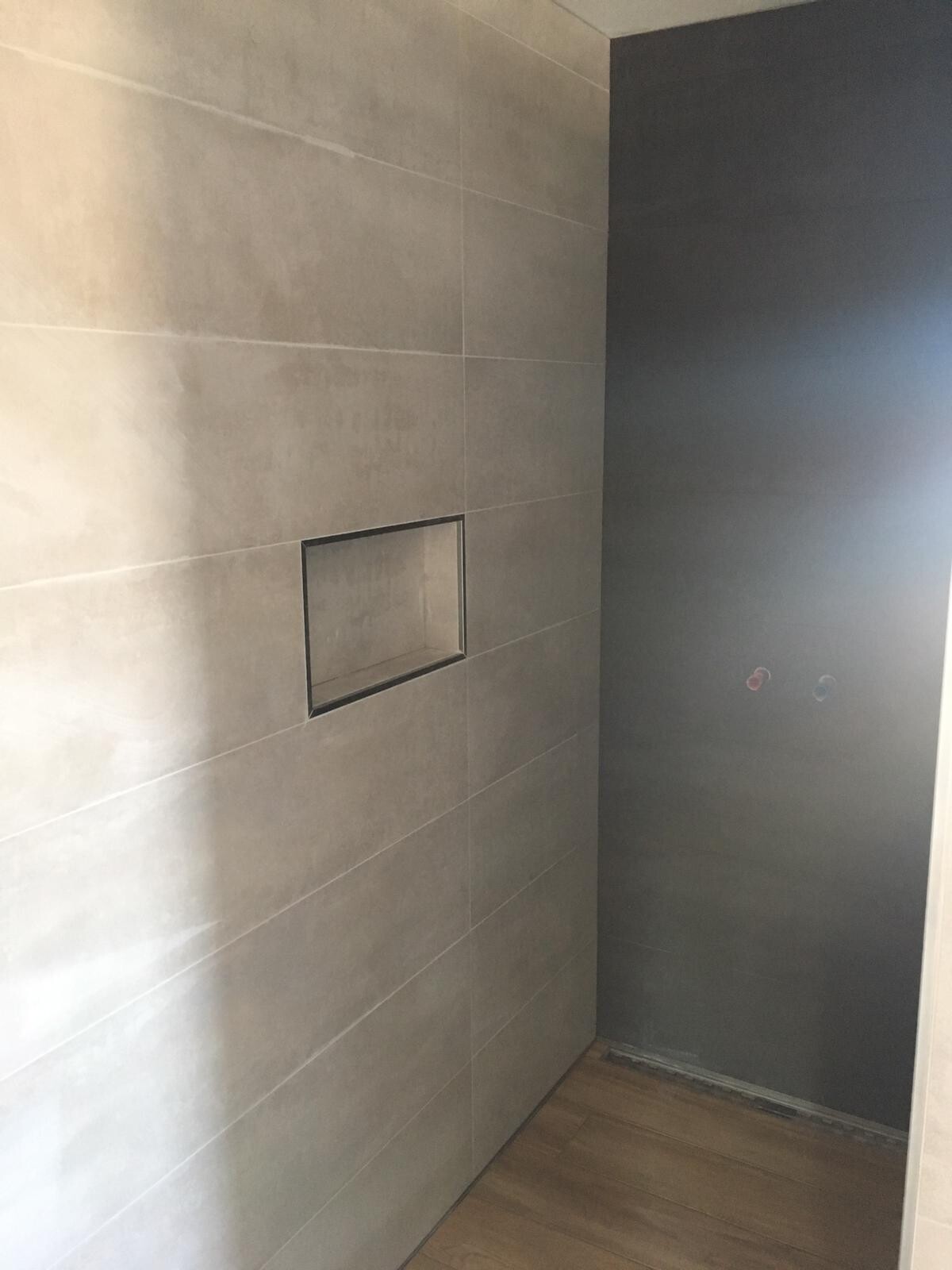 Dusche mit grauen Wandfliesen, linke Nische, dunkle Wand rechts, Wasseranschlüsse, Holzboden.