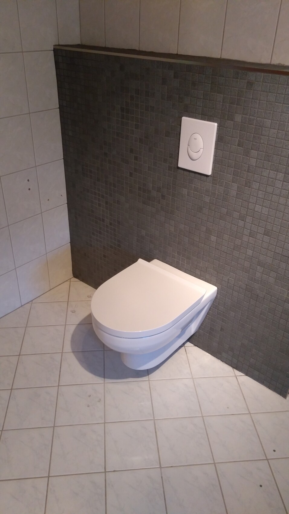 Weiße wandmontierte Toilette vor dunkelgrauer Mosaikwand mit Spülplatte