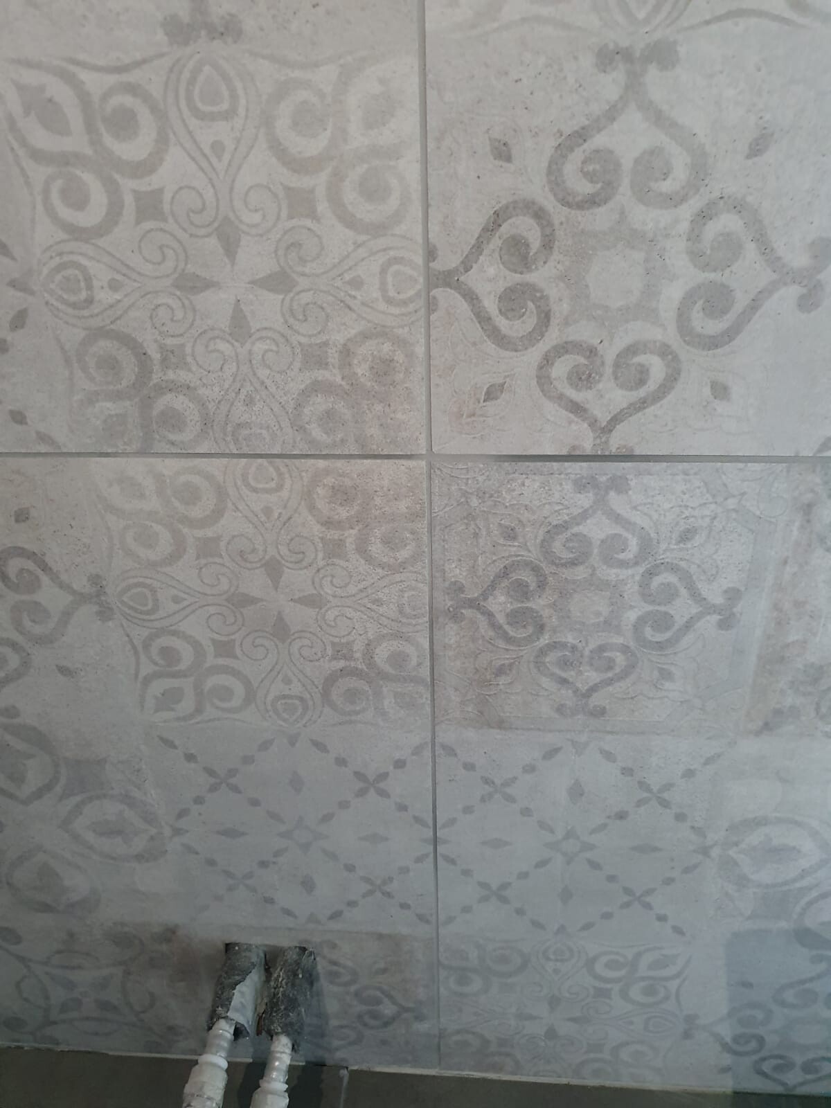 Graue Wandfliesen mit ornamentalem Muster; unten links sichtbare Wasserrohre.