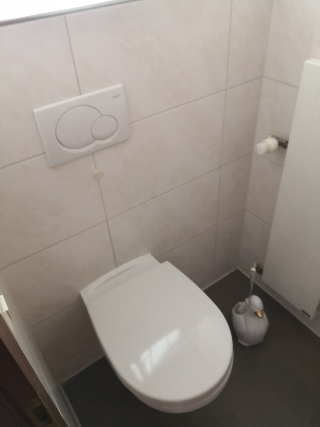 WC mit Wandspülkasten, graue Fliesen, geschlossener Toilettensitz, Bürste rechts.
