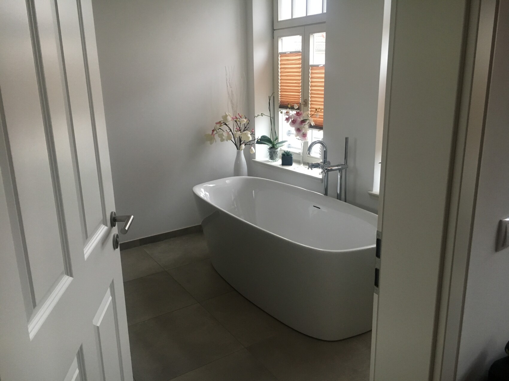 Freistehende weiße Badewanne vor Fenster mit Holzjalousien und Orchideen im Badezimmer