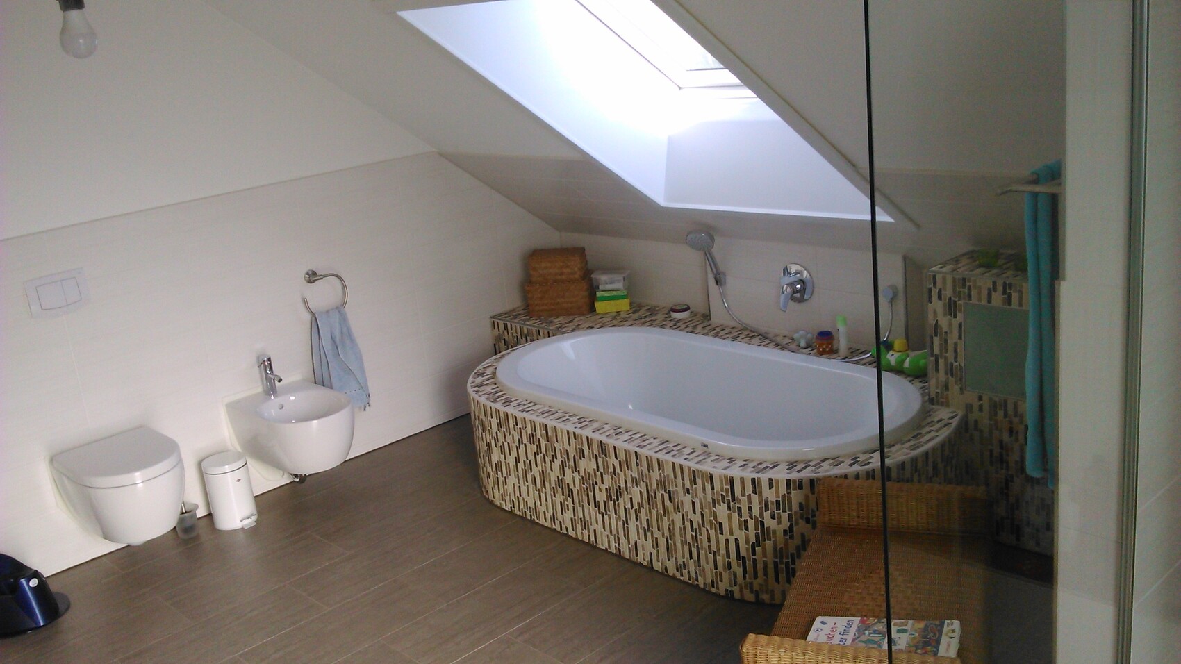 Helles Badezimmer mit ovale Badewanne in Mosaikverkleidung, Dachfenster über der Wanne und Toilette.