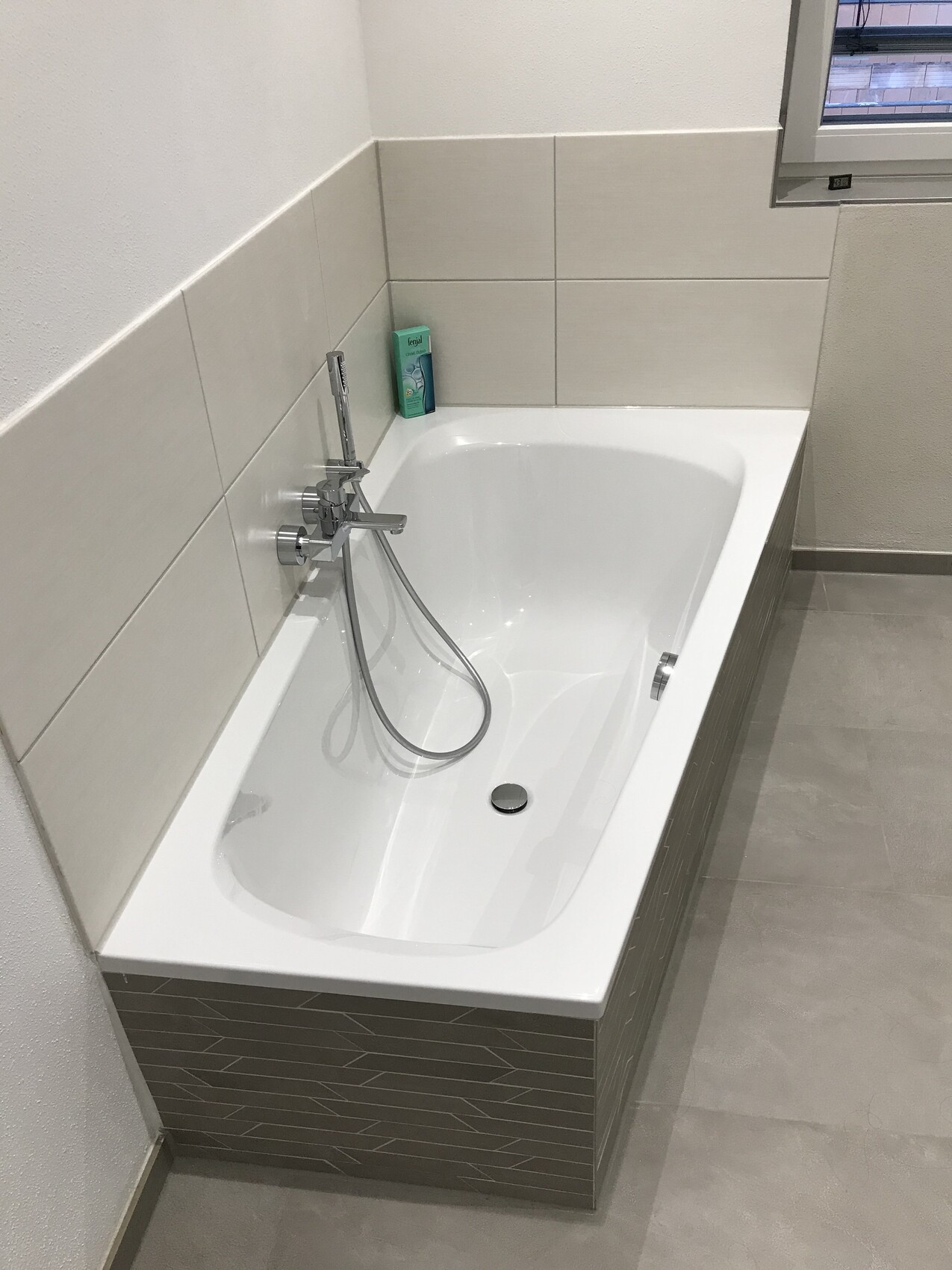 Weiße Badewanne mit Dusche und Handbrause, grüne Seifenflasche am Rand.