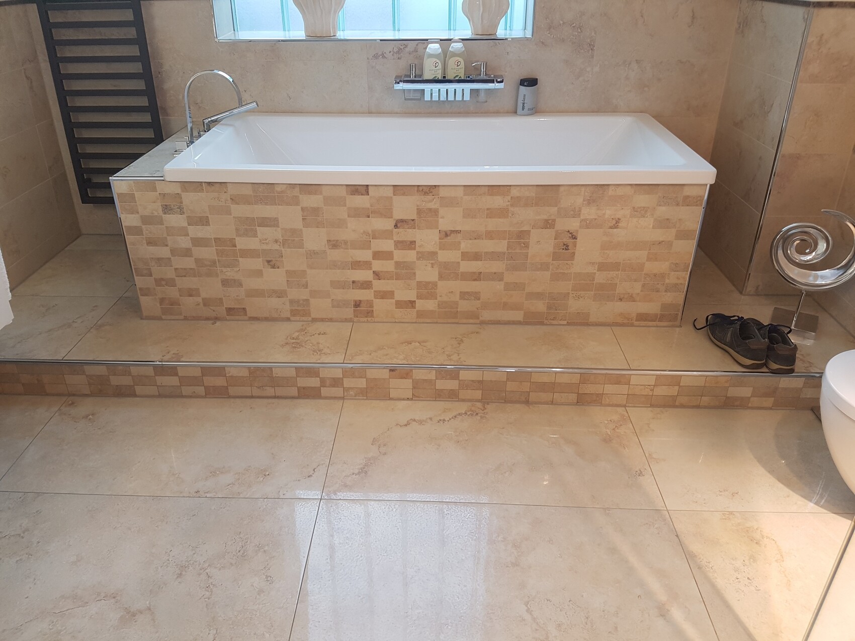 Große Badewanne mit beige Mosaikfliesen, Fenster dahinter; Schuhpaar neben spiralförmigem Hocker.