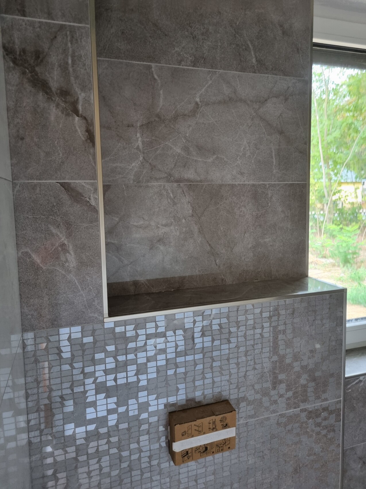 Graue Fliesenwand mit Mosaikbereich unten; kleines Kartonpaket klebt an der Wand.