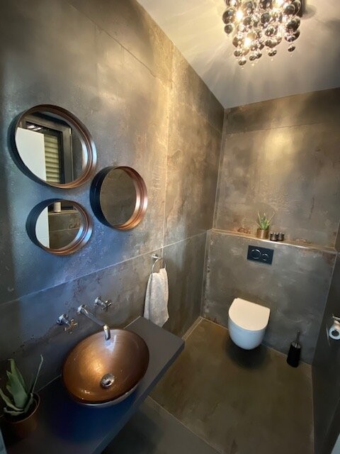 Dunkles Badezimmer mit kupferner Becken-Schale, drei runde Spiegel, Wandtoilette und Kronleuchter.