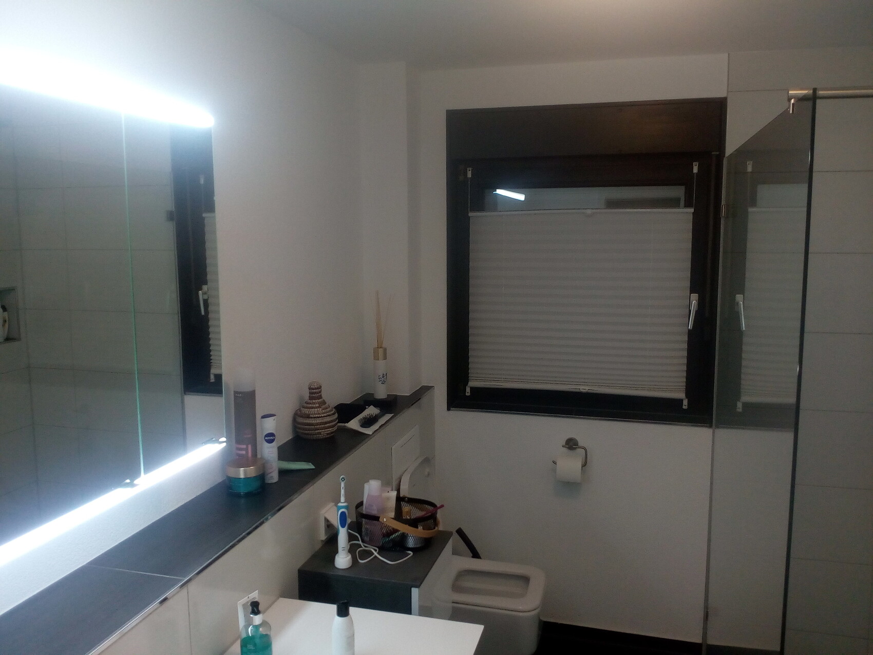 Badezimmer mit Waschbecken, Spiegel, Ablage, Fenster mit Rollladen, Dusche.