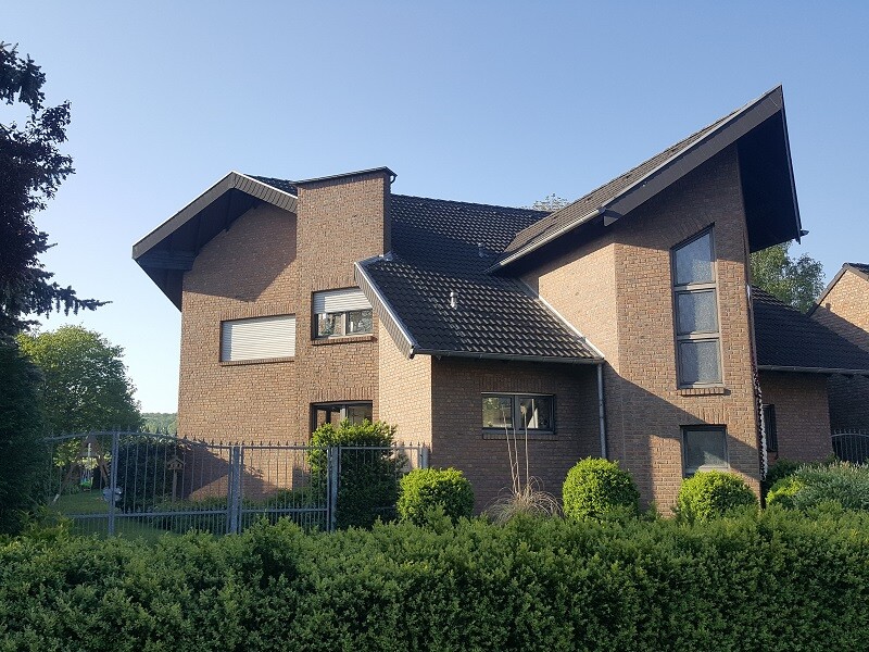 Zweistöckiges Backsteinhaus mit dunklem Ziegeldach, Fensterfronten und Gartenzaun.
