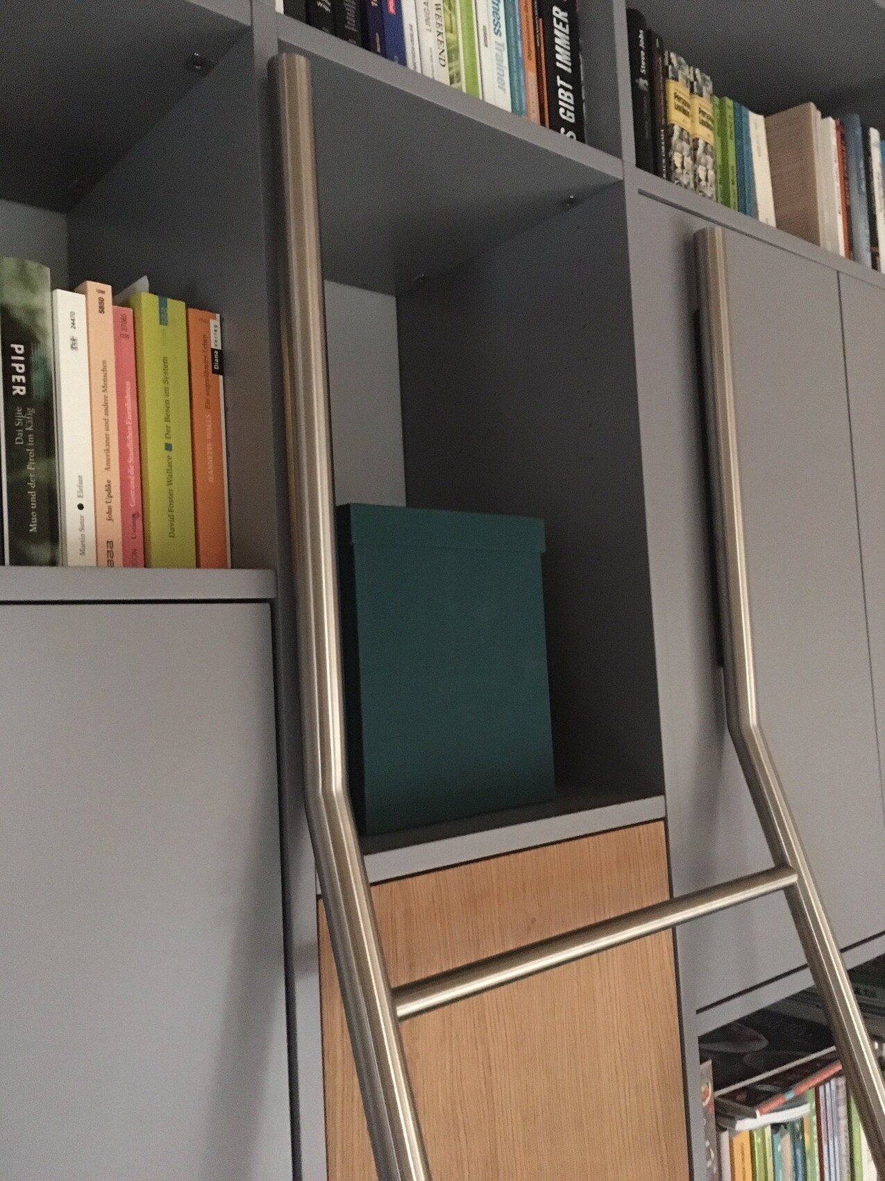Grauer Wand-Schrank mit offener Regalfläche, Metallleiter, grüne Box und Bücher.