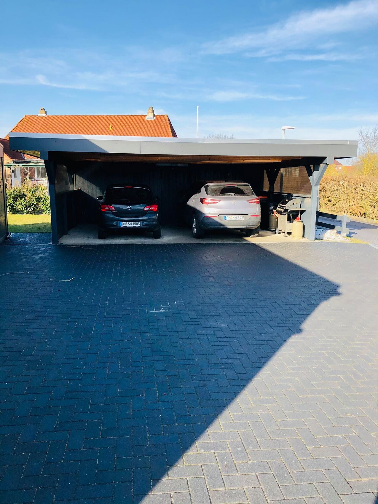 Doppelcarport mit zwei Autos, Grill und Propanflasche vor Häuserzeile, blauer Himmel.