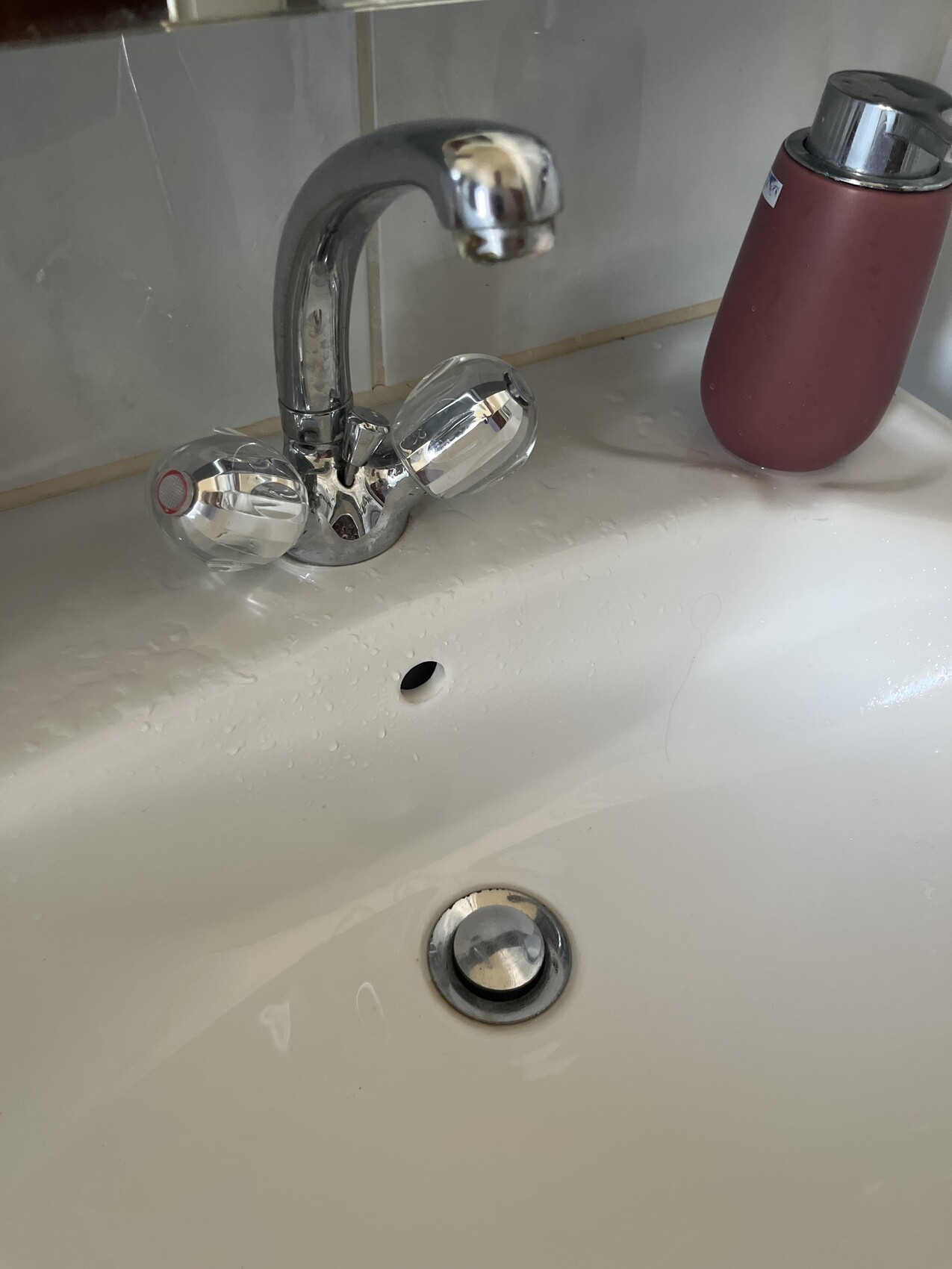 Waschbecken mit glänzendem Chrom-Wasserhahn und zwei Griffen; rosa Seifenspenderflasche daneben.