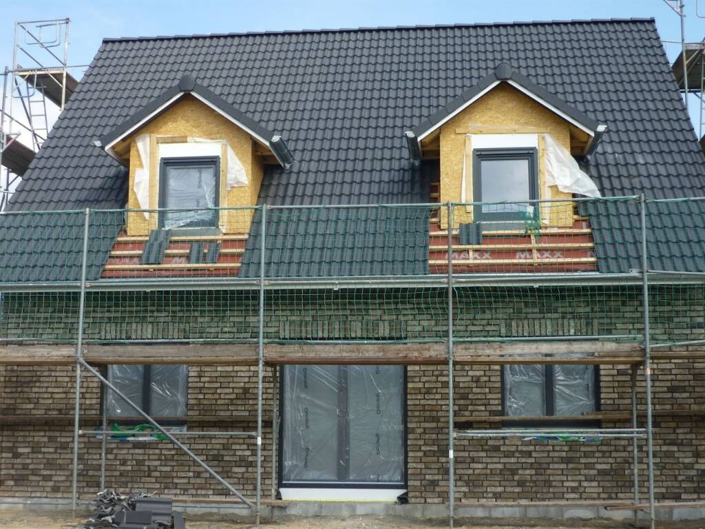 Haus im Bau mit zwei Dachgauben, dunklen Ziegeln, Gerüst und Baufolie an Fenstern