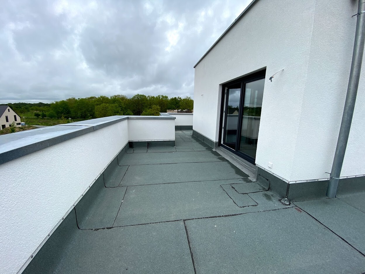 Dachterrasse eines weißen Gebäudes mit Glas-Schiebetür, grauem Boden, Blick auf Bäume
