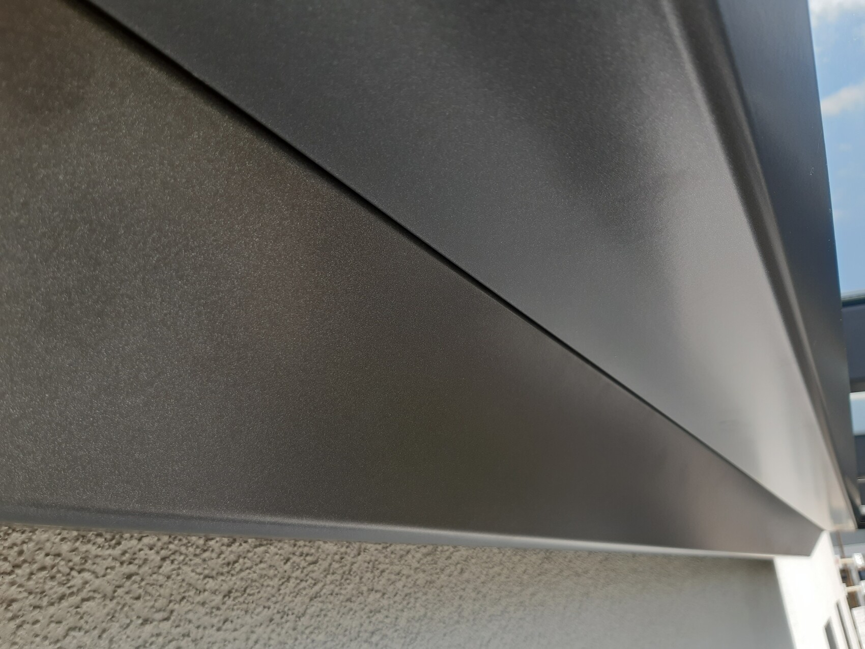 Nahaufnahme grauer Metallverkleidung mit diagonalem Fugenverlauf über rauer Wand, Himmel sichtbar.