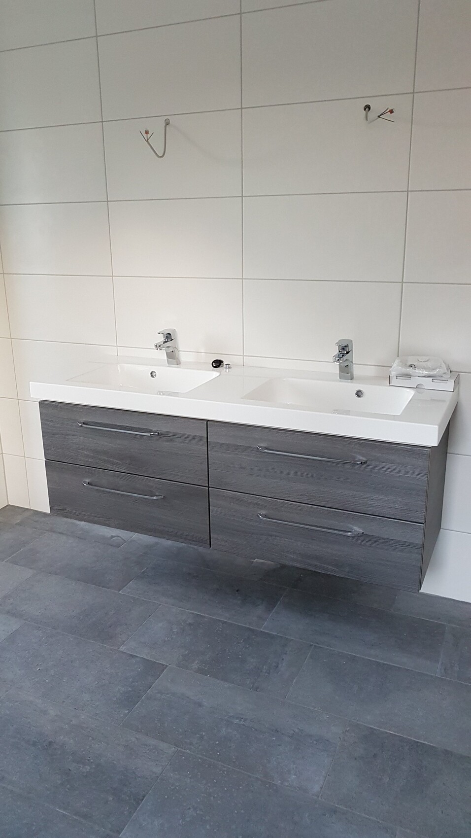 Doppelwaschbecken mit grauem Unterschrank und zwei Wasserhähne vor weißen Wandfliesen.