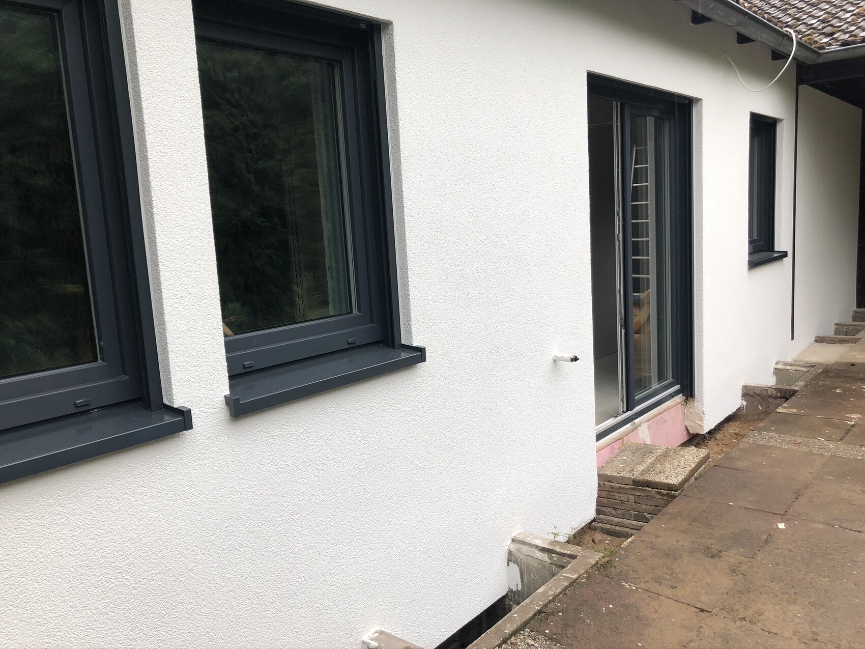 Weiße verputzte Hauswand mit dunkelgrauen Fensterrahmen und Glastür; breiter Steinweg davor.