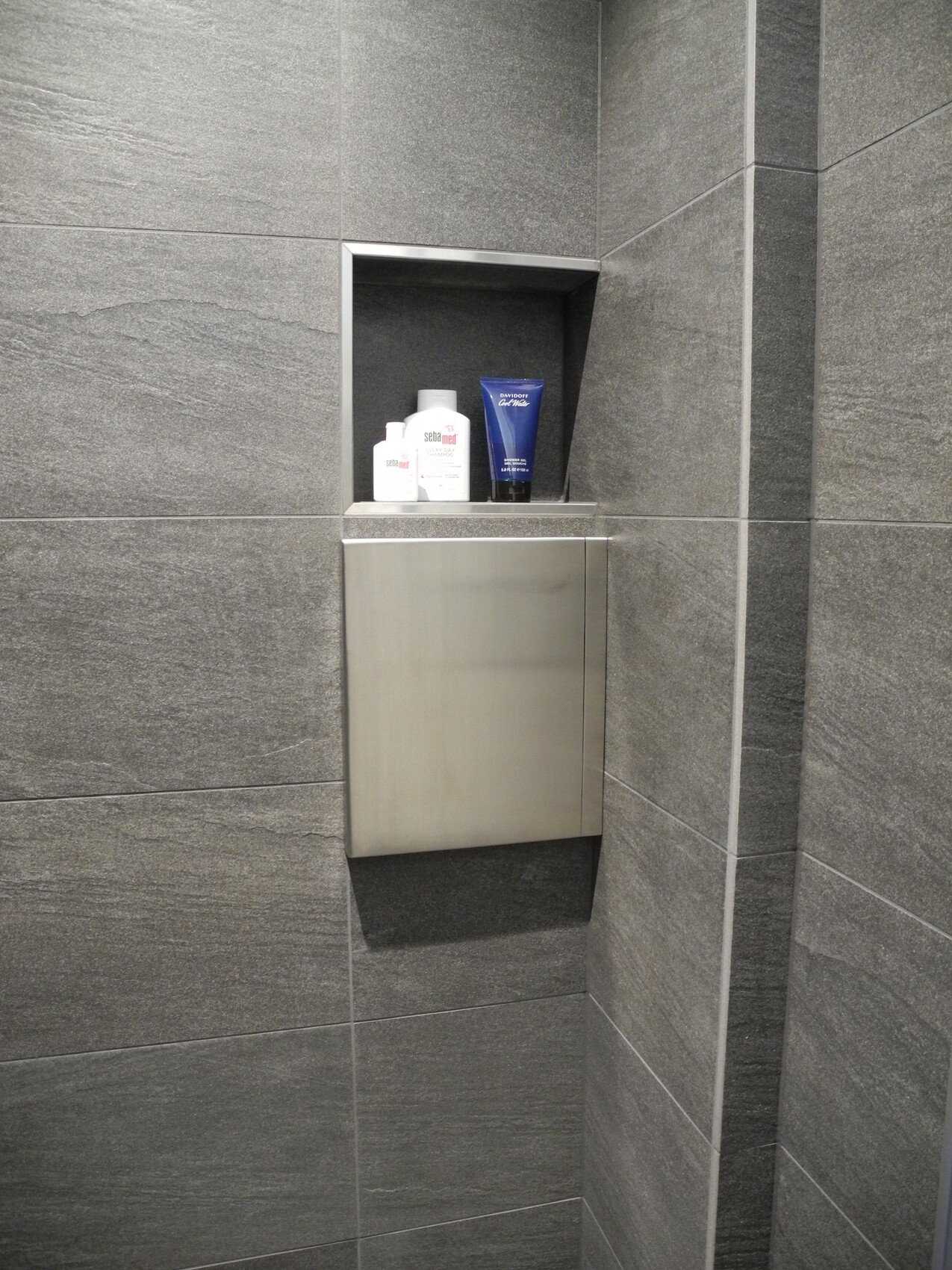 Nische in grau-schieferwand in Dusche mit Shampoo-Flaschen und einer blauen Tube auf Metallablage.