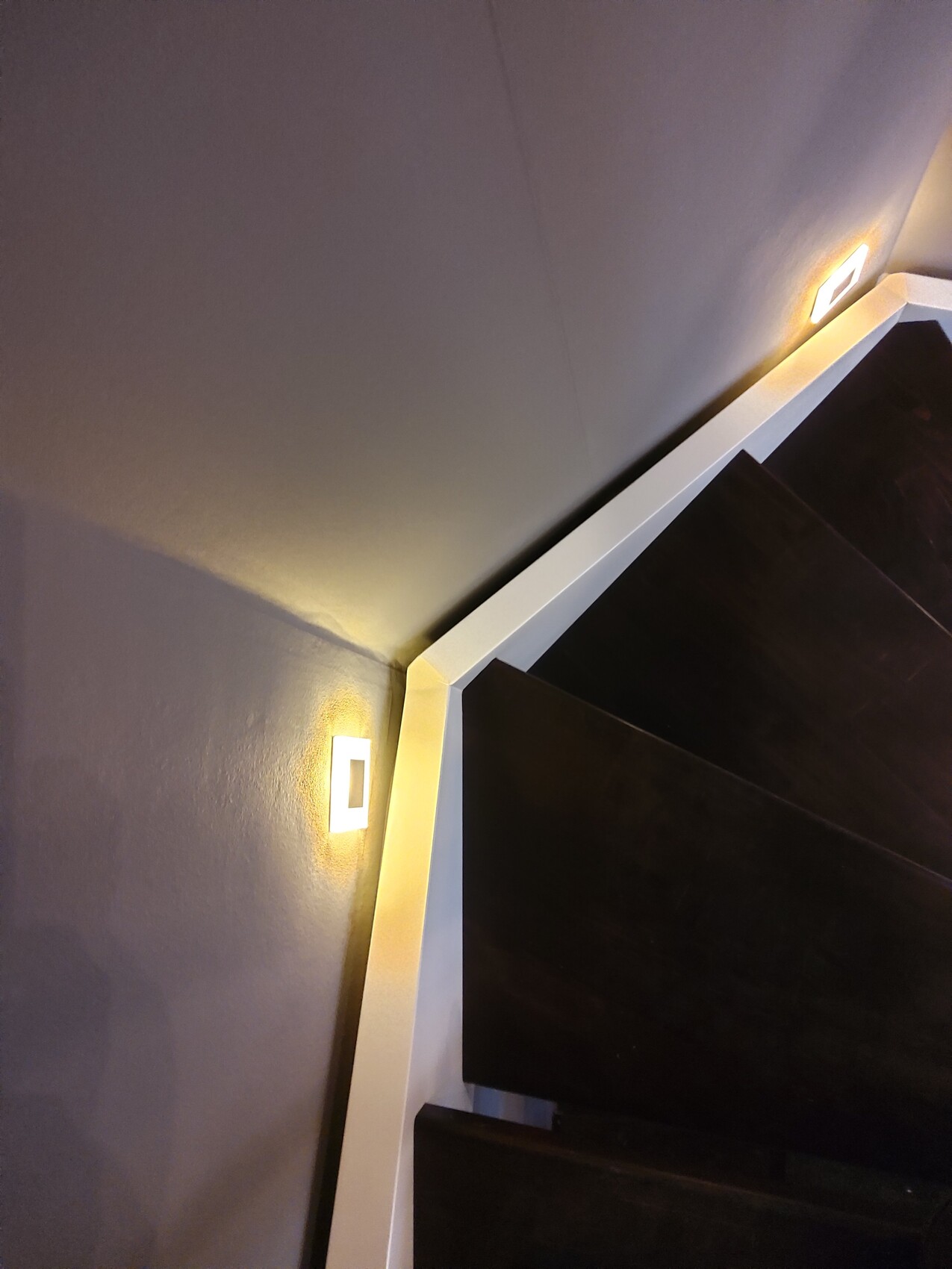 Moderne dunkle Holztreppe an grauer Wand, weiße Treppenkante und warme Wandlampen.