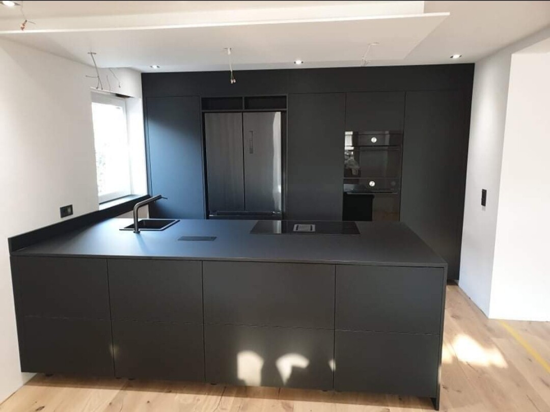 Moderne dunkle Küchenzeile mit Insel, Spüle, Backofen und Kühlschrank im hellen Raum.