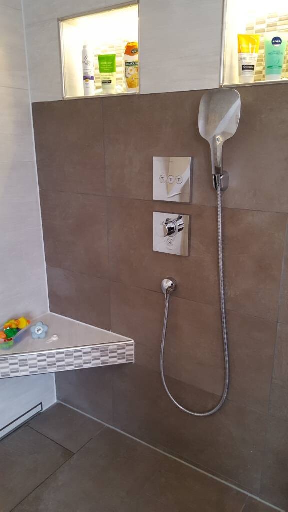 Dusche im Badezimmer mit Handbrause, grauen Fliesen und Ablage.