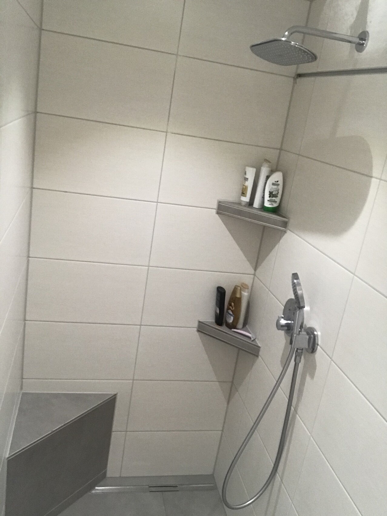 Moderne Dusche mit Regenkopf, Handbrause und zwei Ablagen mit Pflegeprodukten an weißen Fliesen.