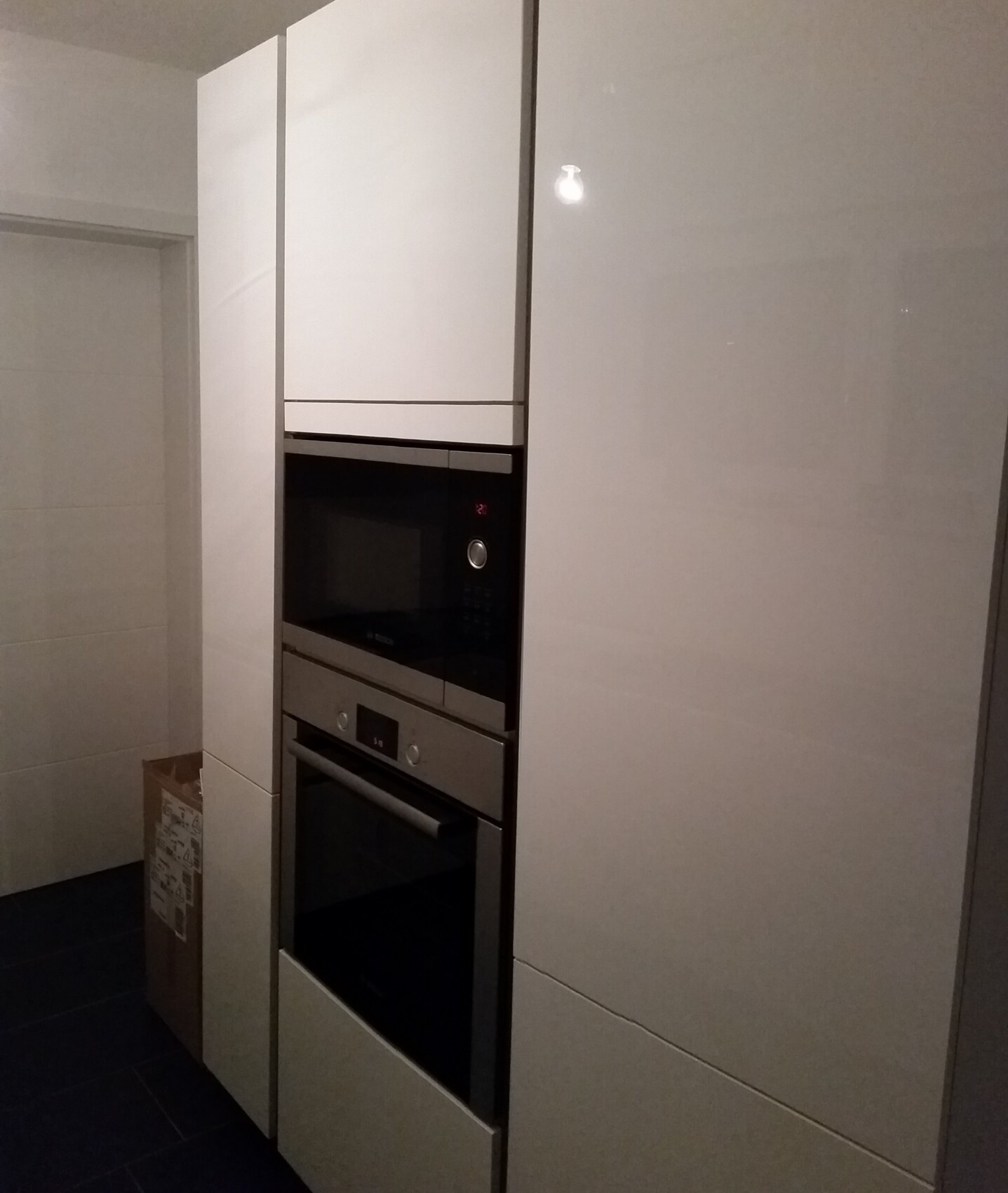 Moderne weiße Küchenzeile mit Hochschrank; integrierte Mikrowelle über Backofen, Karton links.