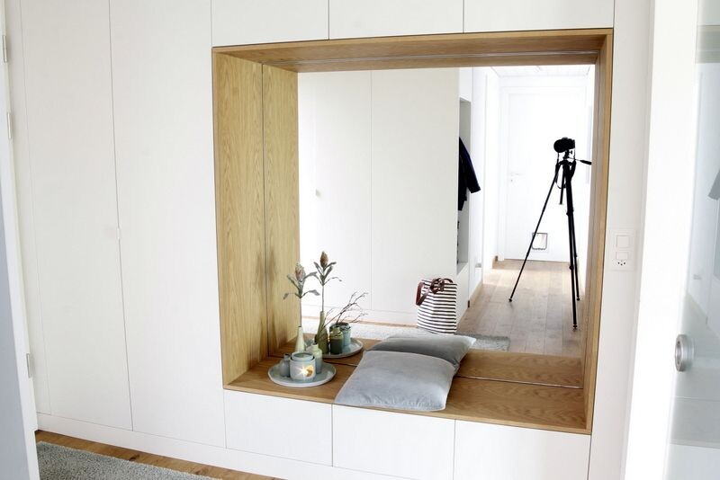 Weiße Einbauwand mit Holzrahmen, Sitzbank mit Kissen, Pflanze und Kerzen; Kamera-Stativ im Hintergrund.