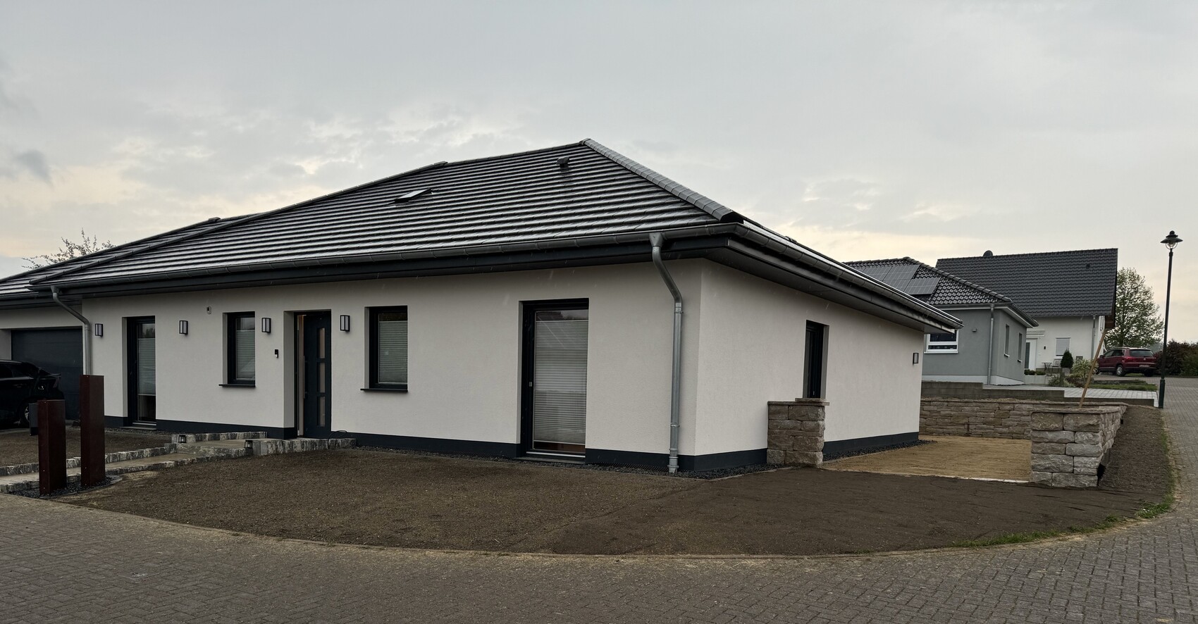 Modernes, einstöckiges Haus mit weißer Fassade, dunklem Ziegeldach, Eingangstür und Fenstern, Vorgarten.