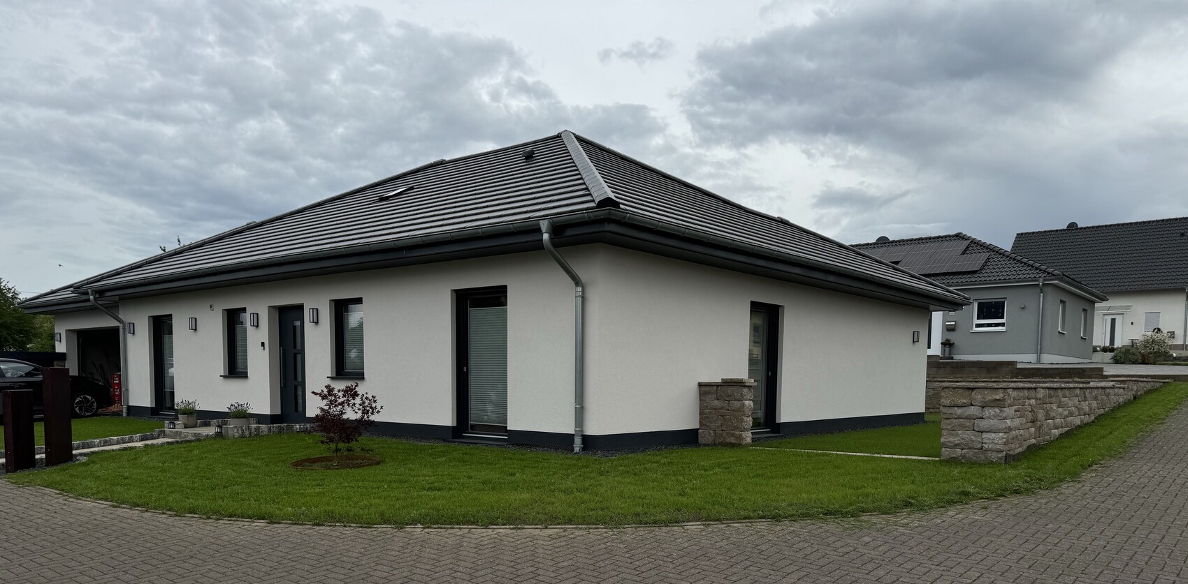 Modernes Einfamilienhaus mit grauem Dach, weißer Fassade, grünem Garten und Steinmauer.