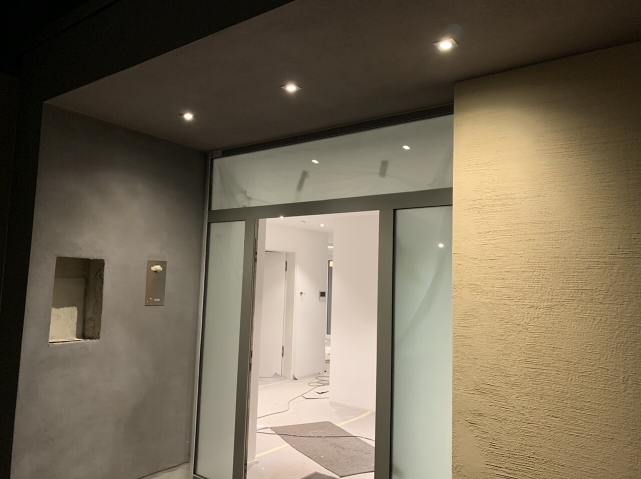 Glastür-Eingang zu hellem Innenraum; linke Wand mit Nische, Kabel auf dem Boden.