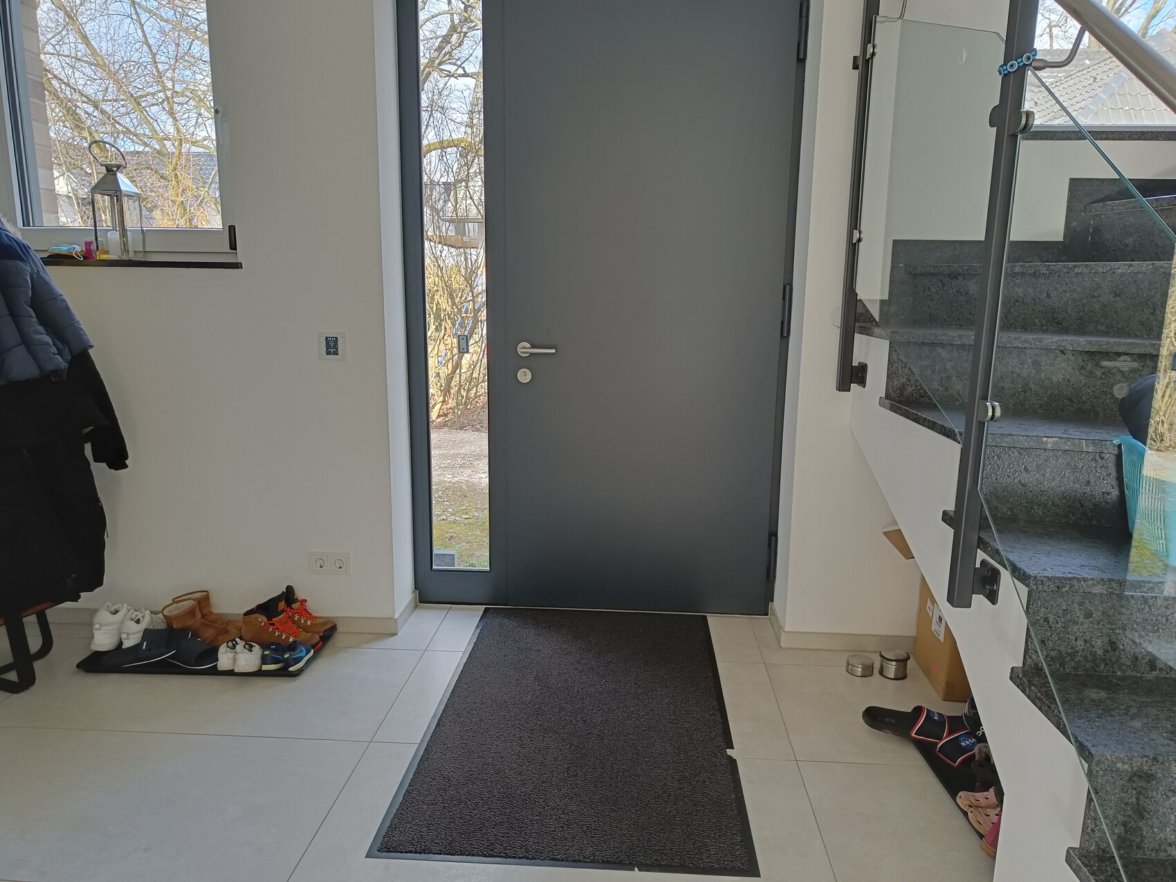 Eingangsbereich: dunkle Tür, Schuhpaare auf Matte, Treppe mit Glasgeländer rechts.