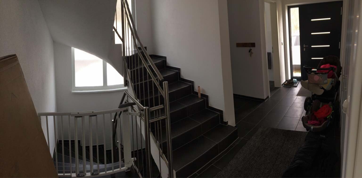 Innenflur mit Treppe, Metallgeländer, heller Fensterbereich, Eingangstür und Kinderwagen.
