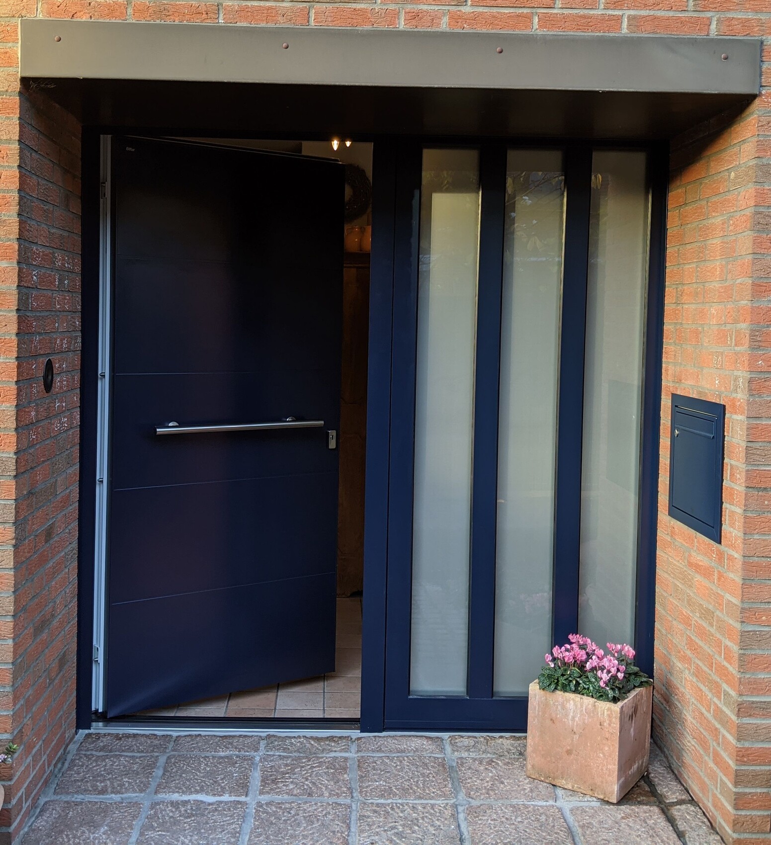 Offene dunkelblaue Haustür mit Glasfenstern; Backsteinwand und Pflanzkübel mit pinken Blumen rechts.